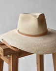 A Ninakuru Ember hat with a broad brim and a leather band rests on a wooden stool against a plain, light gray background.