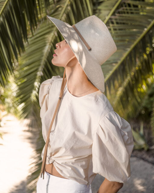 A woman in a light-colored outfit and a wide-brimmed Ninakuru Ember hat stands under palm leaves, looking up thoughtfully.