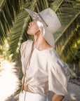 A woman in a light-colored outfit and a wide-brimmed Ninakuru Ember hat stands under palm leaves, looking up thoughtfully.