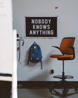 An office with a humble vibe features an orange chair, a blue backpack on the wall, and the Oxford Pennant "Nobody Knows Anything" Camp Flag in black and white.