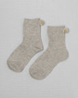 Unified’s Winter Pom Pom Socks feature a light gray crew design with small beige pompom accents at the back, displayed flat to highlight their subtle natural hues.