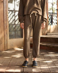 A person stands on wooden steps wearing Hey Gang's The Sweatpants in brown, paired with a matching sweatshirt and black slip-on clogs. Sunlight casts shadows on the porch and door behind them, with their upper head out of frame.