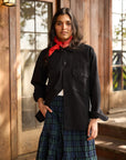 A woman with long dark hair, wearing a black button-up shirt and a blue-green plaid skirt, stands before a wooden building. Around her neck, she wears the Hey Gang HG Bandana—an everyday accessory. She gazes at the camera with a neutral expression.