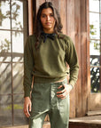 A woman stands before wooden doors, sunlight streaming in behind her, wearing The Sweatshirt in olive green by Hey Gang and matching pants, accessorized with a black bandana around her neck.