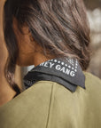 A person with long, wavy brown hair wears an olive green top and The HG Bandana by Hey Gang around their neck, which features "HEY GANG" in white letters. The image highlights the back and side of their head and shoulders.