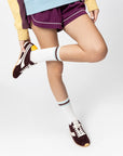 A person wears the ONCEPT Essex Sneaker, a blue and yellow sweatshirt, purple pinstripe shorts, and white socks with blue stripes, standing with one leg bent and crossed over the other against a white background.