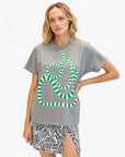 A woman with wavy blonde hair wears Clare Vivier's Original Tee Snake, a 100% cotton gray T-shirt with a green and white striped snake graphic, paired with a high-slit black-and-white patterned skirt against a white background.