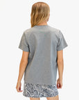 A person with long, light brown hair is shown from the back, wearing a Clare Vivier Original Tee Snake gray 100% cotton t-shirt and black-and-white patterned shorts, standing against a plain white background.