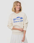 A woman with blonde hair smiles in the Clare Vivier Oversized Sweatshirt S25—cream fleece cotton with a blue fish graphic and "Liberez Les Sardines"—paired with a white skirt. The look is an instant conversation starter against a white backdrop.