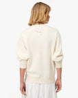 A person with short blonde hair is seen from behind wearing a Clare Vivier Oversized Sweatshirt S25 in off-white fleece cotton and a white skirt, with blue "Liberez Les Sardines" lettering on the upper back.