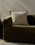 The PJBR0001 Natural GO Pillow by Loloi Rugs, featuring cream and beige stripes with brown piping and a hidden zipper, rests on an olive green velvet sofa beside a black side table topped with a minimalist vase of bare branches.