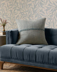 The PCJ0018 Blue Natural sofa by Loloi Rugs features tufted blue polyester, wooden legs, and a large woven cotton pillow with a blue-green stripe. It sits beside leafy wallpaper and an antique bronze side table with a vase of flowers.