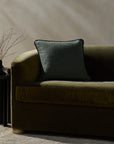 A dark green velvet sofa with a Loloi Rugs Hudson Blue Pillow 20x20 sits next to a small black side table holding a vase with thin branches. Sunlight casts soft shadows on the beige wall behind.