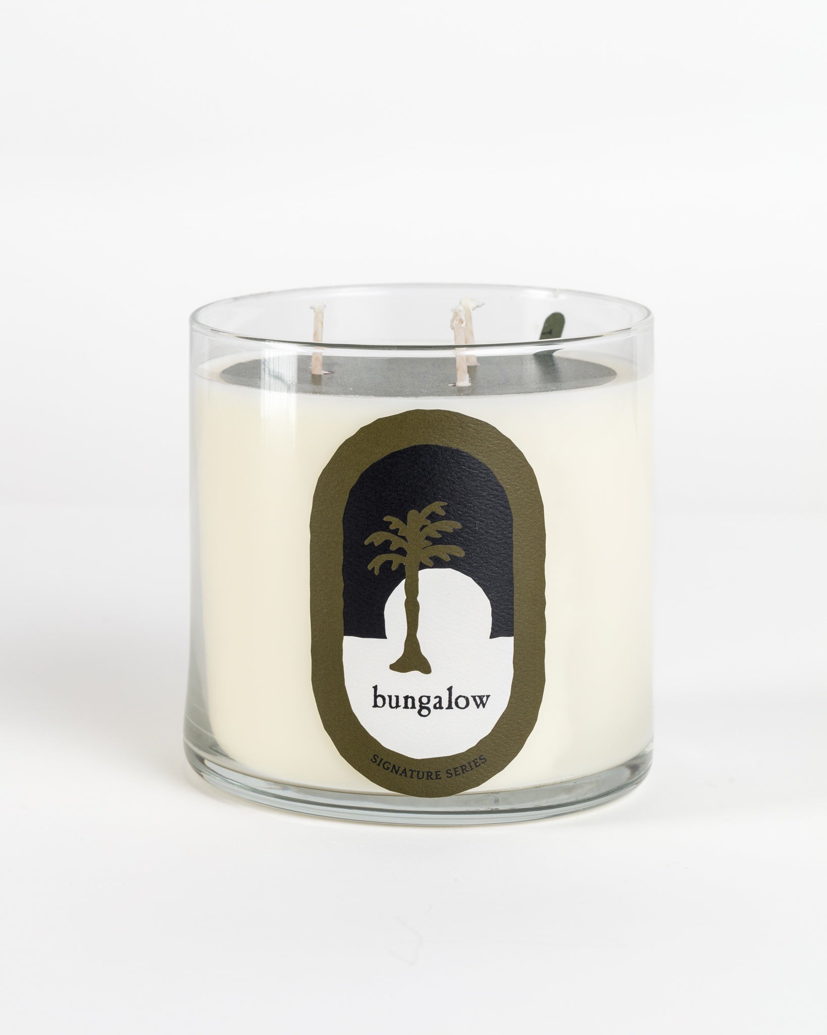 The New Signature Candle by Mame, infused with sandalwood, features a white candle in a glass jar with two wicks. Its label showcases a black and white design with a palm tree silhouette and the word "bungalow" in a simple font, all set against a plain white background.
