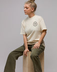 A woman with blonde hair in a bun sits on a wooden block, wearing the Things Unseen T-Shirt by Things Unseen with a small chest logo and olive green pants, gazing calmly to the side.