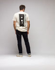 A man stands facing away in the Things Unseen T-Shirt by Things Unseen, featuring a vintage cassette design inspired by the Beastie Boys' License to Ill album. He pairs it with dark jeans and white shoes, set against a plain light gray background.