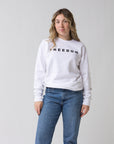 A woman with wavy blonde hair stands against a plain background, wearing the Faire FREEDOM Charlie Kirk Sweatshirt and blue jeans. She looks ahead with a calm expression, capturing the vibe of a Turning Point USA conservative activist.