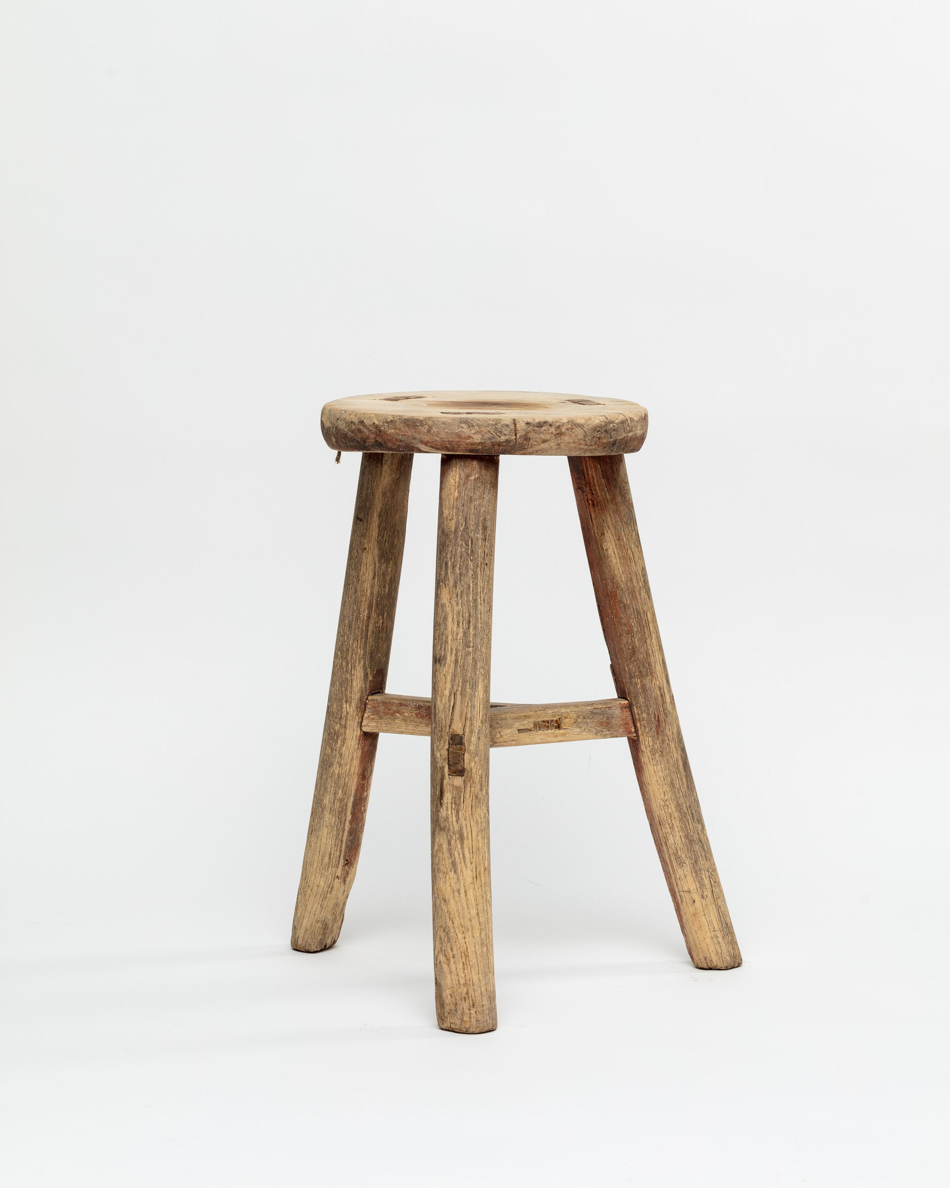 The ROUND STOOL by Indus Design Imports is a weathered, rustic wooden stool featuring four legs and a round seat. Showcasing timeless elegance, the stool's surface reveals natural wood texture and imperfections, giving it a worn, antique appearance. This piece of artisan craftsmanship stands against a plain white background.