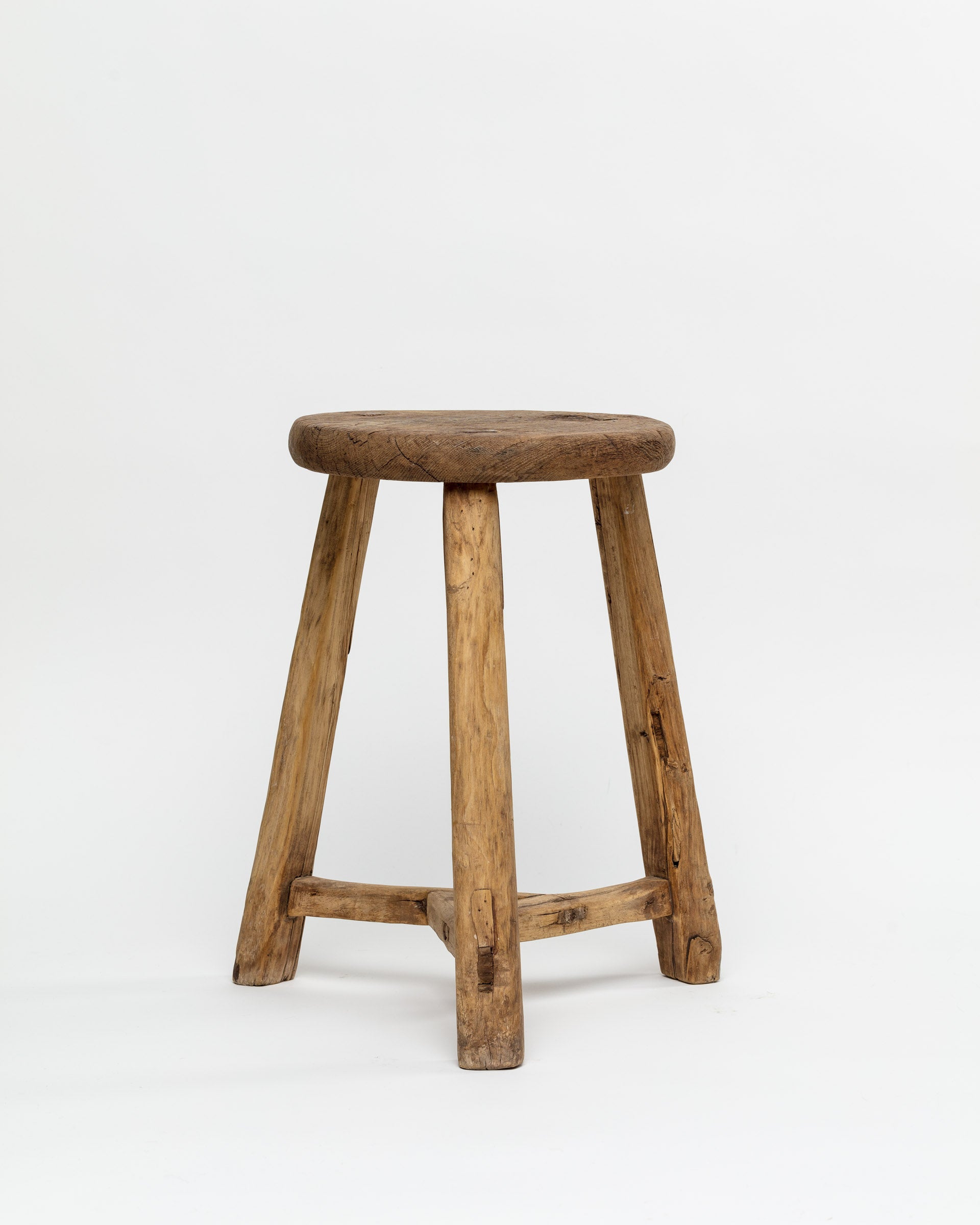 A ROUND STOOL 48 from Indus Design Imports, with a round top and three legs, sourced from Scottsdale, Arizona, set against a plain white background. The wood has a weathered appearance, indicating age or frequent use.