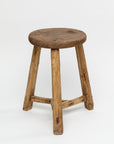A ROUND STOOL 48 by Indus Design Imports with a round seat and three splayed legs, showcasing visible aged wood textures and grain against a plain white background in a Scottsdale bungalow.