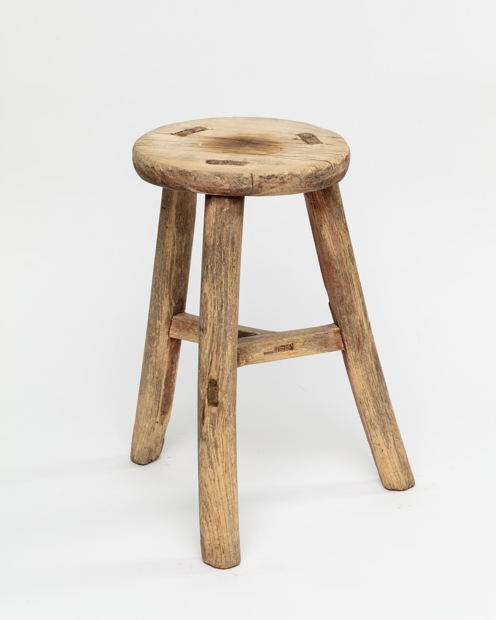 Introducing the ROUND STOOL by Indus Design Imports, a rustic piece crafted from wood with three legs and an unfinished, rough surface. Showcasing artisan craftsmanship, this stool features visible grain patterns and natural imperfections in the wood, giving it a weathered and vintage appearance. It stands alone against a plain white background.