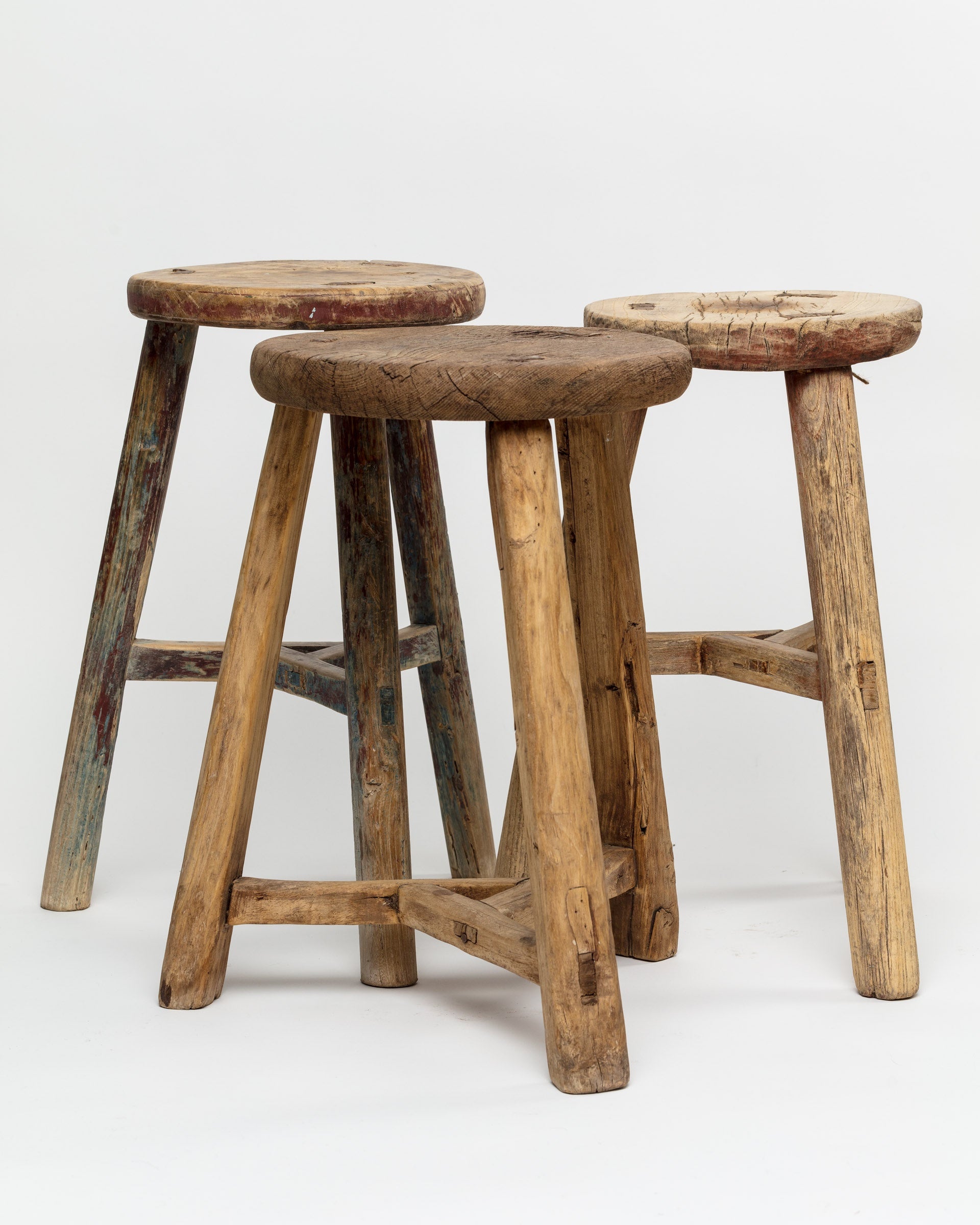 Three ROUND STOOLs by Indus Design Imports, each of different heights, are arranged closely together against a plain white background. These imported handmade vintage stools feature weathered surfaces with visible grain and wear, highlighting their artisan craftsmanship and imparting a timeless elegance.