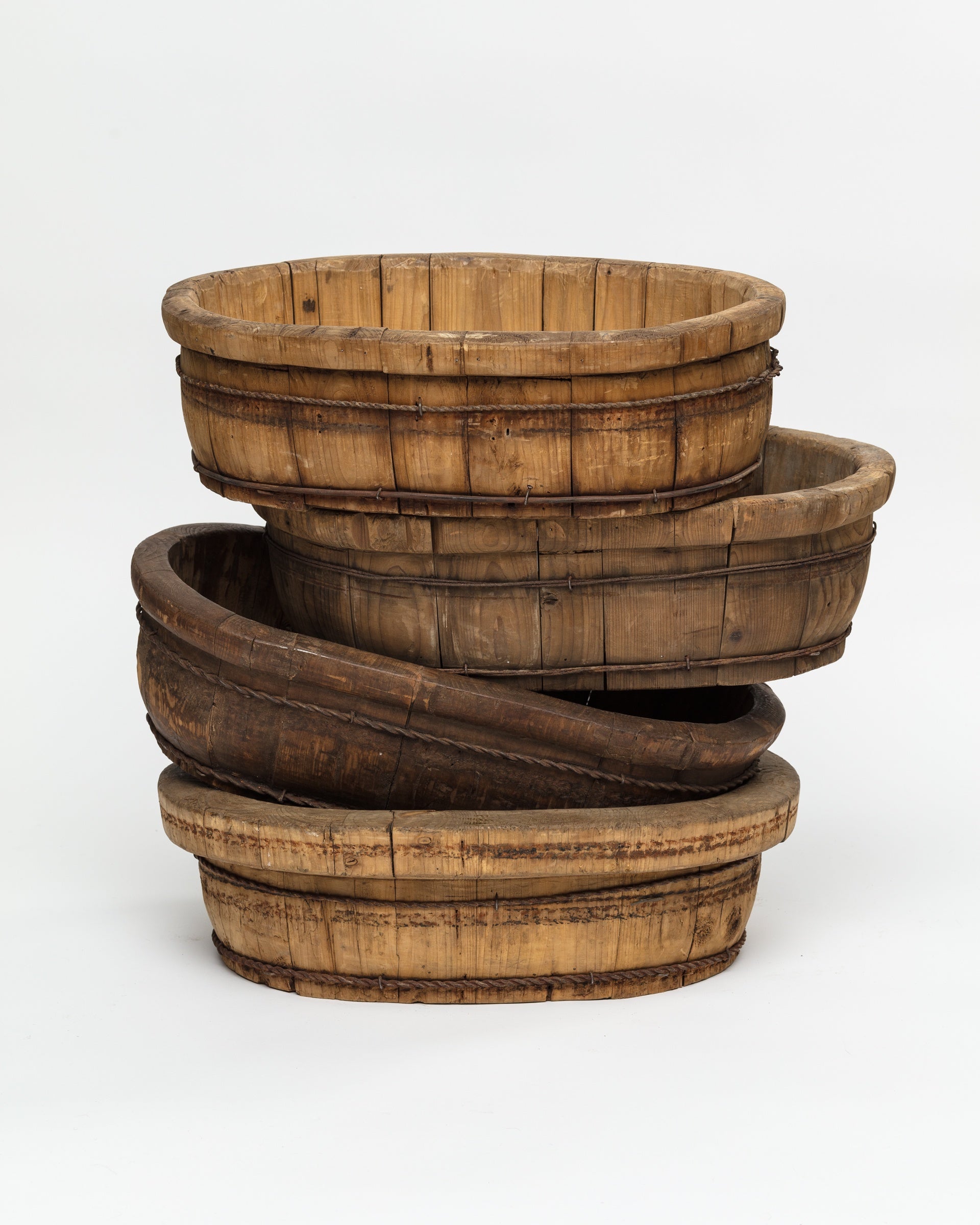 A stack of four rustic, round wooden baskets of varying sizes exudes rustic elegance. The handmade vintage baskets are slightly worn, displaying visible wood grain and metal bands that highlight their aged charm. They nest inside one another, creating a tiered arrangement against a plain white background. These are the Indus Design Imports Oval Wood Bucket 10.
