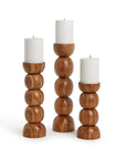 The PDT001 Pillar Candleholders by Two's Company feature hand-crafted, stacked rounded shapes in natural wood finish, each holding a white pillar candle. Three varying heights are arranged side by side on a plain background.