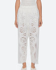 A person wearing Sea New York Gardener Pants—vintage-inspired sheer white pants with floral cut-out embroidery and an elastic waistband—paired with white flats stands against a plain light background.