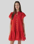 A woman smiles gently while wearing the Sea New York Pippa SS Mini Dress, a vintage-inspired bright red mini dress with lace details, flutter sleeves, and front button-down design, standing against a plain light gray background.