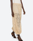 A person wears the Sharleen Lace Pant by Sea New York—sheer beige floral lace with a balloon silhouette, elastic waistband, and gathered ankles—paired with a white textured top and black heeled sandals.