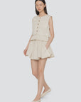 A woman with straight long hair stands against a plain background wearing the Sea New York Angelina Suiting Flare Short, a sleeveless beige button-up top, and matching beige flats. The shorts feature pleats and an elasticized waistband.