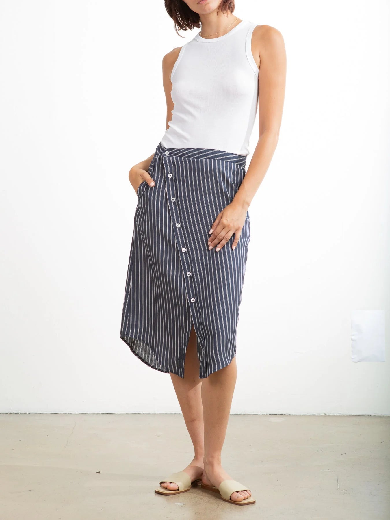A woman stands against a white background wearing a sleeveless white top, beige slide sandals, and the Pharaoh Nico Skirt—a blue and white vertical striped 100% cotton button-front skirt with front pockets. Her left hand rests in her skirt pocket.