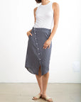 A woman stands against a white background wearing a sleeveless white top, beige slide sandals, and the Pharaoh Nico Skirt—a blue and white vertical striped 100% cotton button-front skirt with front pockets. Her left hand rests in her skirt pocket.