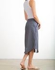 A woman faces away, wearing a white sleeveless top and the Pharaoh Nico Skirt—a blue and white vertical striped 100% cotton midi skirt with front patch pockets—paired with beige sandals against a plain light background.