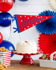 A white frosted cake with cherries sits on a red cake stand, decorated with the “America” Felt Pendant Banner by My Mind's Eye. Red, white, and blue balloons and fans create a patriotic backdrop, with nearby popcorn and strawberries.