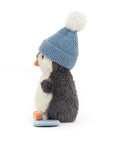 The Jelly Cat Inc. Peanut Penguin Snowboarding plush sits upright in a blue knit hat with white pom-pom, featuring a white belly, gray back, and orange beak and feet, shown in profile on a white background.