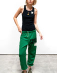 A woman with blonde hair wears a black tank top, green Circa '99 OG Sweat pants by Free City, featuring "FREE CITY" text and the FREECITYLARGE logo, plus green sneakers. She stands on a concrete floor against a plain white background.