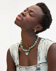 A woman with short curly hair tilts her head back, eyes closed, wearing a sleeveless acid-wash denim top and the Clare Vivier Checker Shell Necklace, along with layered green and silver necklaces, gold stud earrings, against a white background.