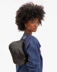 A woman with curly hair in a blue sweater looks over her shoulder, carrying the Clare Vivier Petit Moyen Messenger S25, a black handwoven leather mini crossbody bag. The background is plain white.