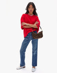 A person with long dark hair in a loose red T-shirt, blue jeans, and white shoes stands confidently holding the Clare Vivier Petit Moyen Messenger S25 with crossbody straps against a plain white background.