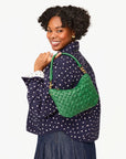 A smiling woman with curly hair, wearing a polka dot jacket and denim skirt, carries the Clare Vivier Petit Moyen Messenger S25, a green handwoven leather purse, over her shoulder.