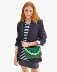A person with long red hair dons a dark blazer over a striped shirt and black skirt, holding the Clare Vivier Shortie Strap Q1—a green bag with vintage brass detailing—against a plain white background.