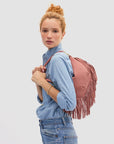 A woman with curly red hair in a bun, wearing a light blue denim shirt and jeans, stands against a plain background with the Clare Vivier Petit Moyen Messenger Suede Fringe bag in mauve suede over her shoulder.
