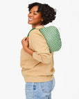 A person with curly hair smiles while holding a Clare Vivier Petit Moyen Messenger S25, a green checkered crossbody bag, over their shoulder. They wear a beige sweatshirt and blue jeans against a plain white background.