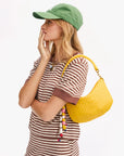 A woman in a green baseball cap and brown striped shirt stands in profile, hand on her chin, carrying the Clare Vivier Petit Moyen Messenger S25—a yellow woven crossbody accented with colorful beads—over her shoulder.