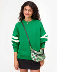 A woman with long curly hair wears a bright green sweatshirt, black mini skirt, necklace, and the Clare Vivier Petit Moyen Messenger S25—a handwoven leather crossbody bag—against a plain white background.