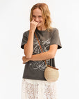 A smiling woman with wavy blonde hair wears a dark graphic t-shirt, a white lace skirt, and carries the Petite Pot de Miel straw bag by Clare Vivier. She stands against a plain light background, one hand gently touching her face.