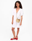 A woman stands against a plain white background wearing a white short-sleeve dress, red shoes, a layered pink necklace, and carrying Clare Vivier's Petite Pot de Miel handwoven straw bag.