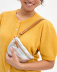 A person wearing a mustard yellow buttoned cardigan smiles while holding a Clare Vivier Shoulder Strap w/Grommets, a small silver crossbody bag featuring brass grommets, gold hardware, and a tan Italian leather strap.